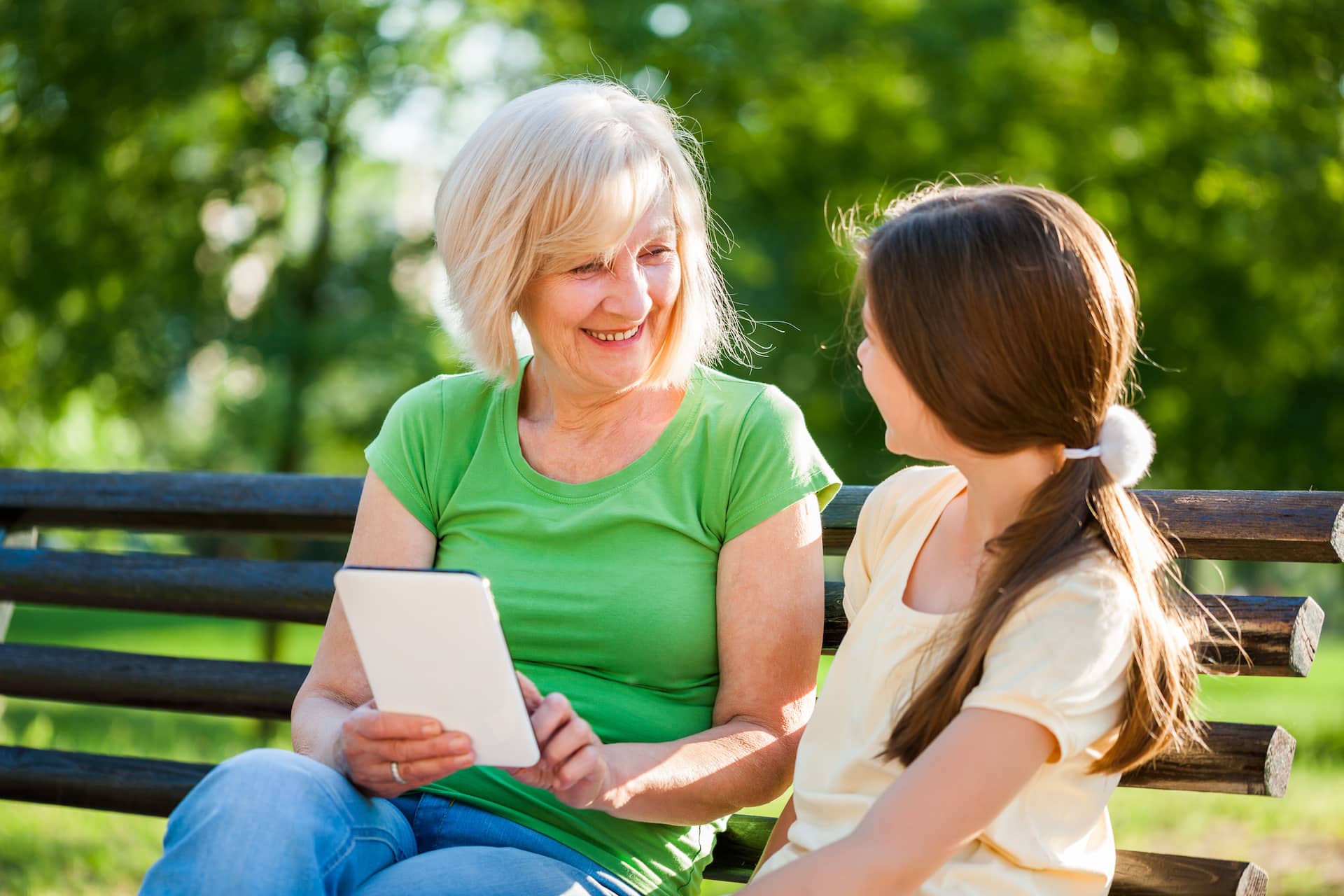 Grandmother and granddaughter are sitting in park and using digital tablet.